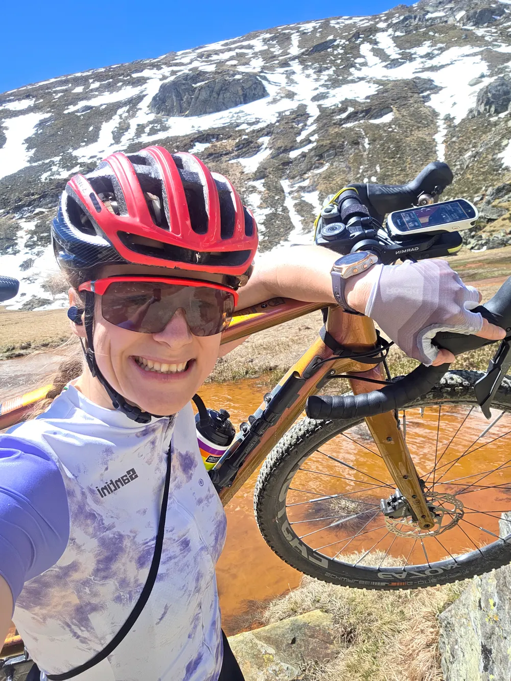 Selfie of Carla in helmet and sunglasses carrying her gravel bike, snowy mountain slopes behind her