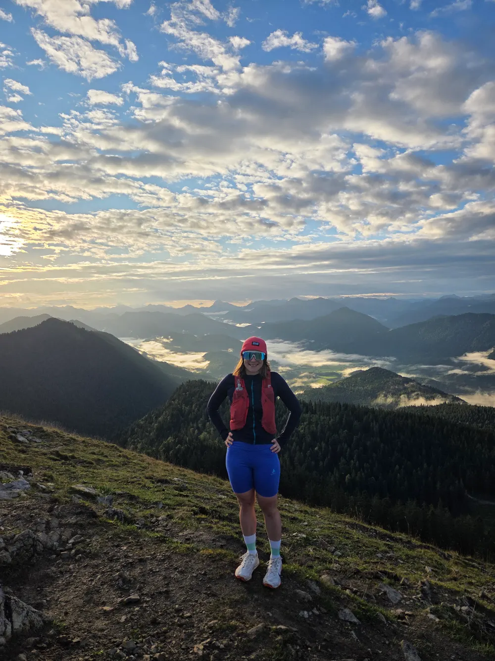 Carla in running kit on a summit at sunrise, with fog filling the valleys of the Bavarian Prealps below