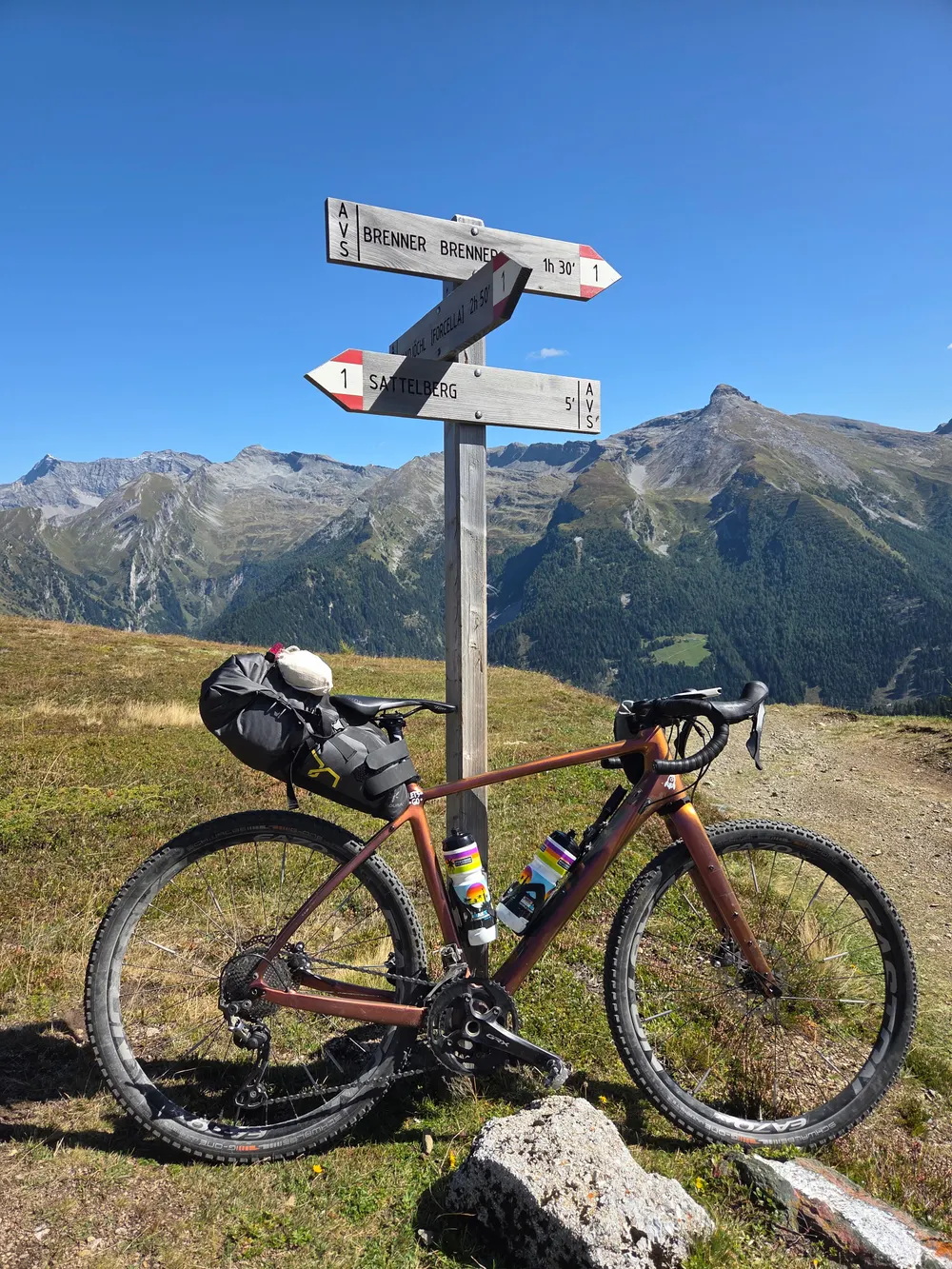 The gravel bike with a saddle bag leaning against a trail signpost towards the Brenner, mountains under a blue sky behind