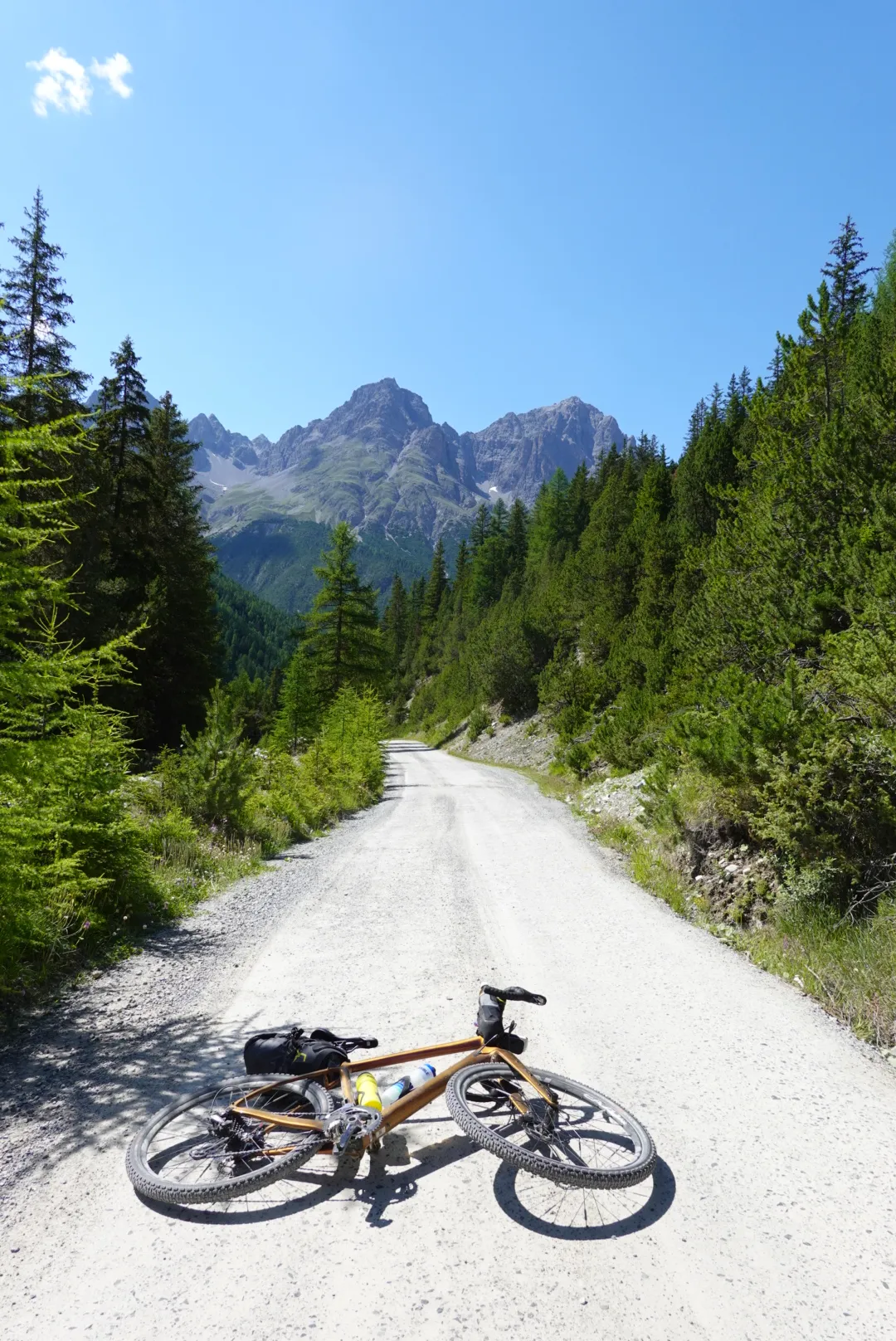 Gravel bike lying on a pale gravel road between spruces, mountain peaks behind
