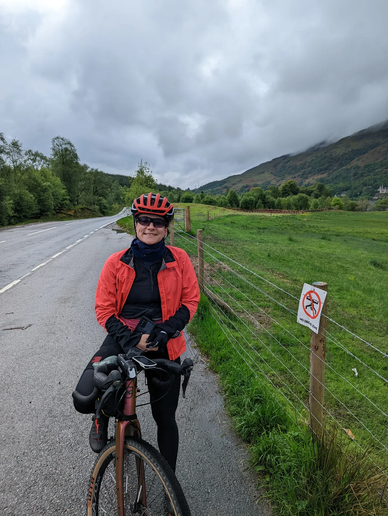 Carla in a red jacket with her bike at the roadside, green hills behind