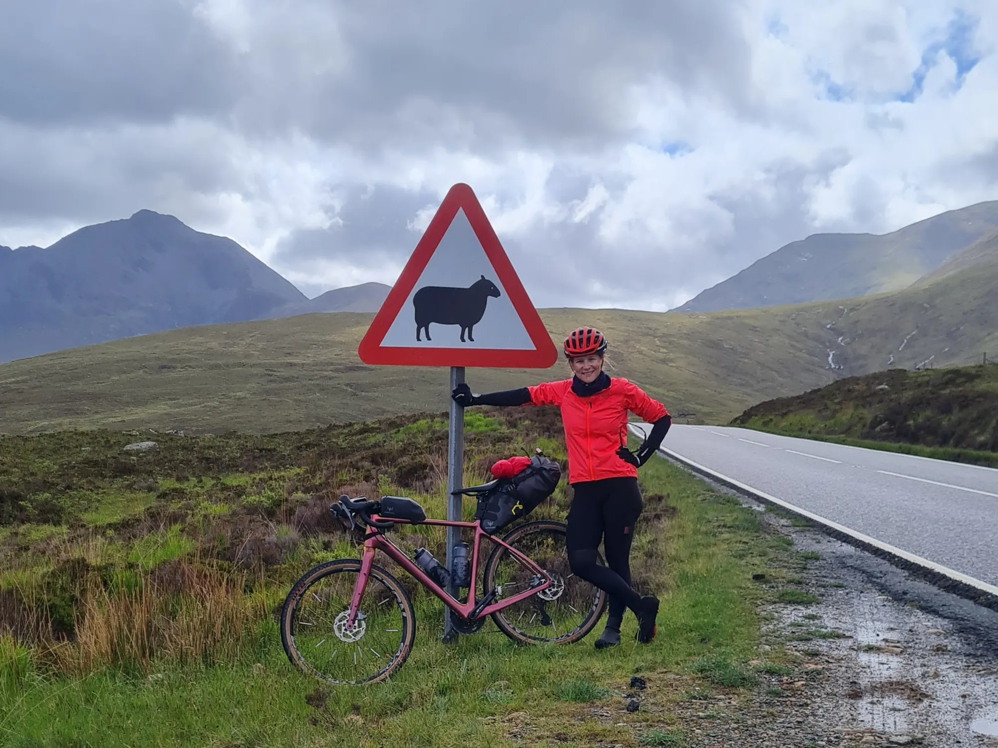 Carla with her bike at a sheep warning sign, dark mountains behind