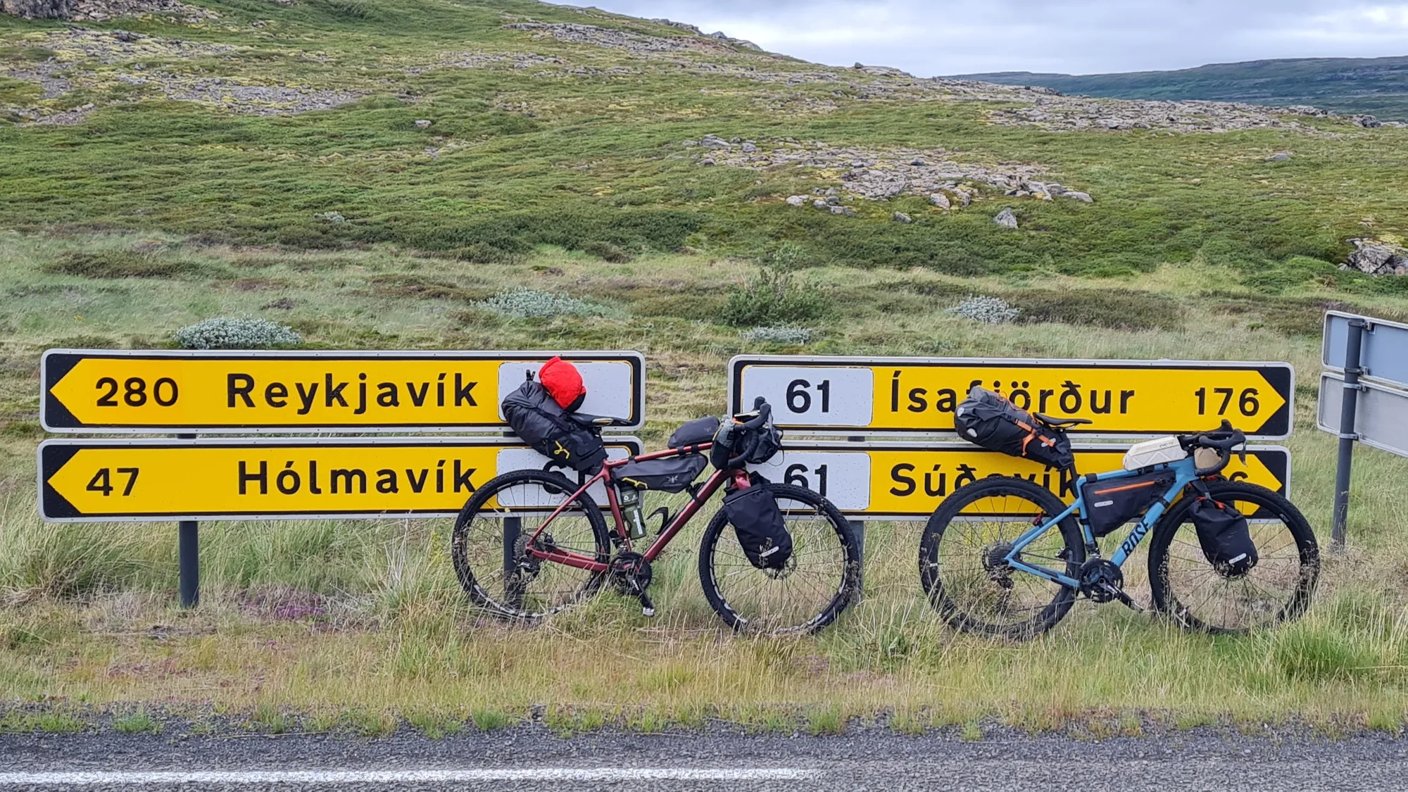 Two loaded gravel bikes in front of yellow road signs to Reykjavík, Hólmavík and Ísafjörður